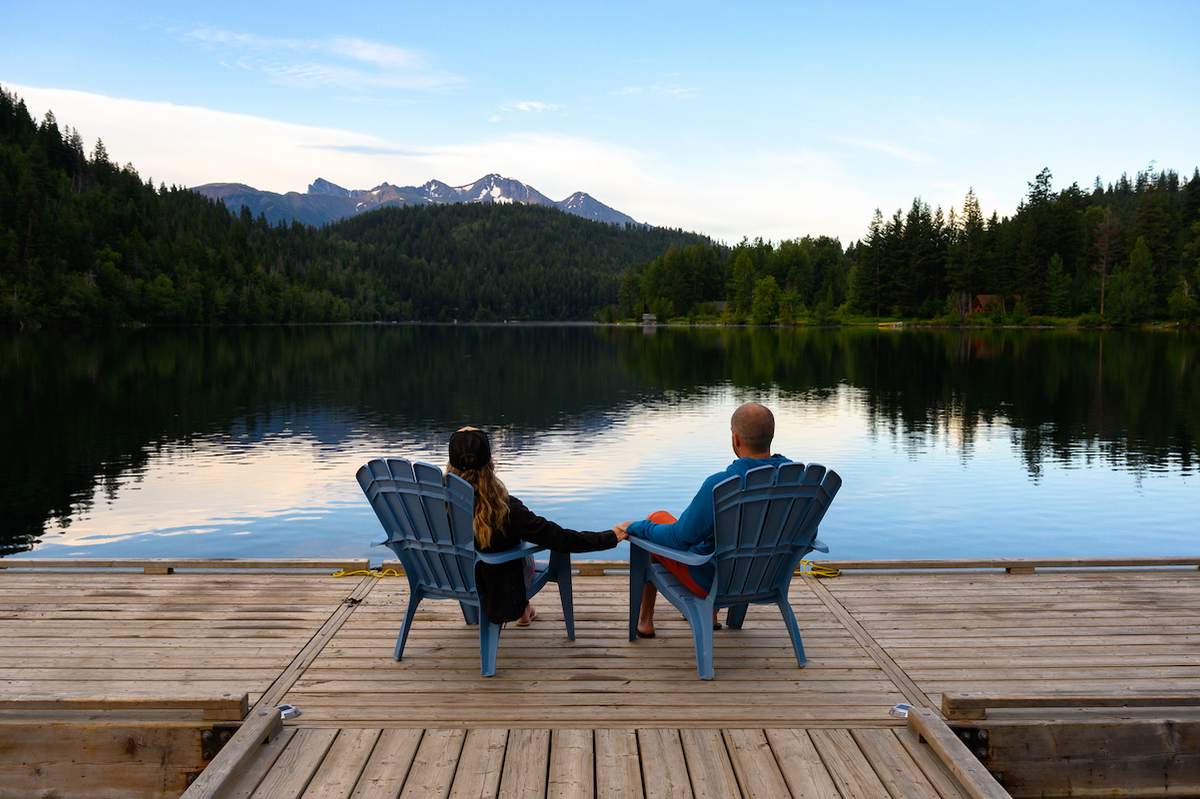 A woman and man sit on chairs on a deck looking at a lake.